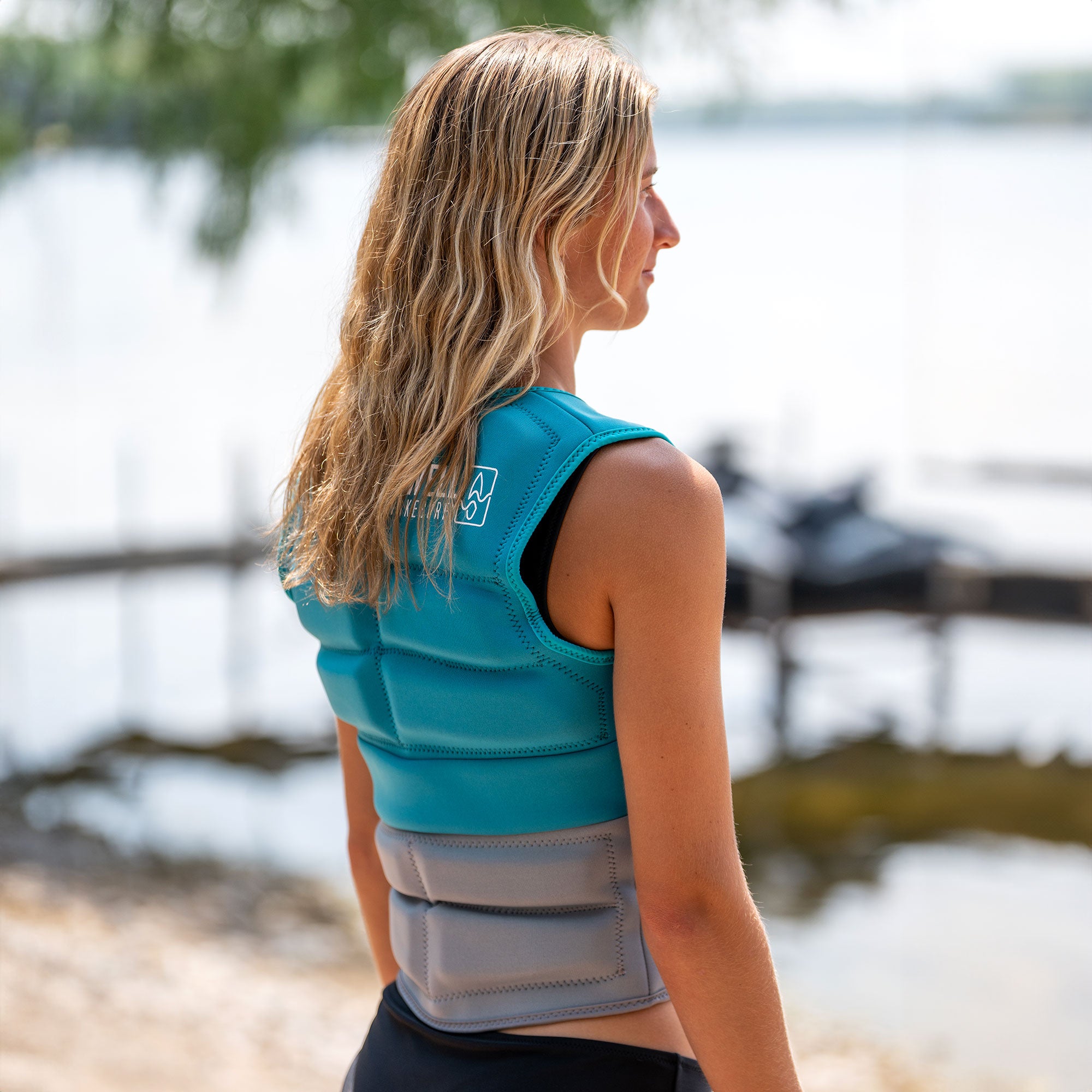 A woman is standing next to a lake in a SWELL Wakesurf Vest - Women's Aqua for comfort and safety.