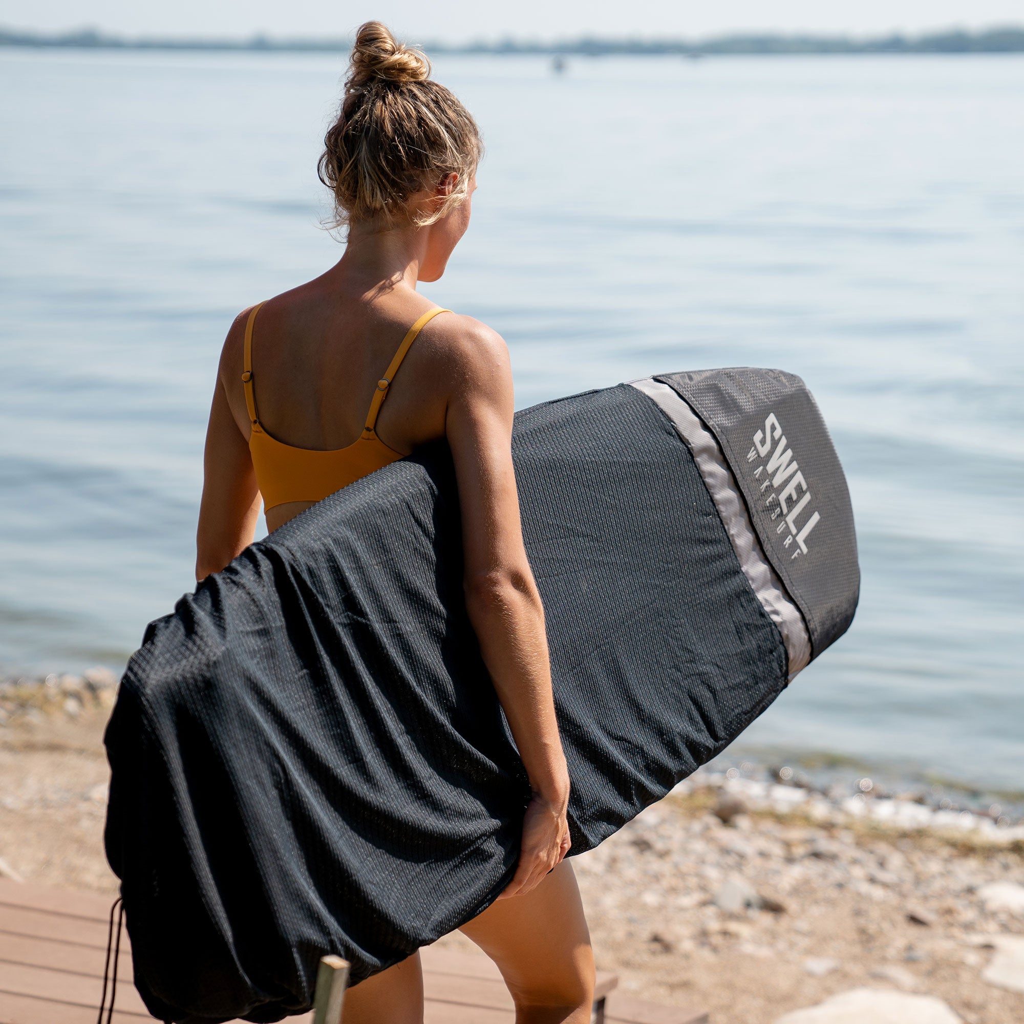 A woman in a bikini carrying a surfboard for UV protection with the SWELL Wakesurf Board Surf Sock - Padded Nose With Nose Pocket.